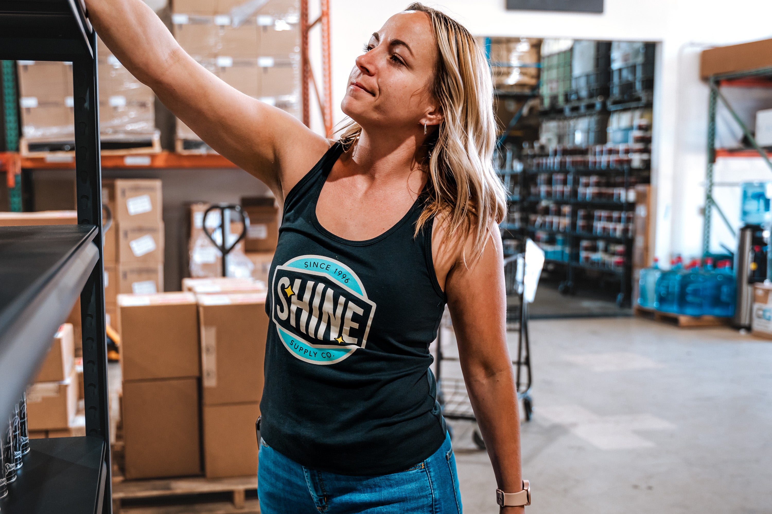 Woman in a warehouse wearing a dark tank top with 'SHINE Supply Co.' logo, reaching for a shelf.