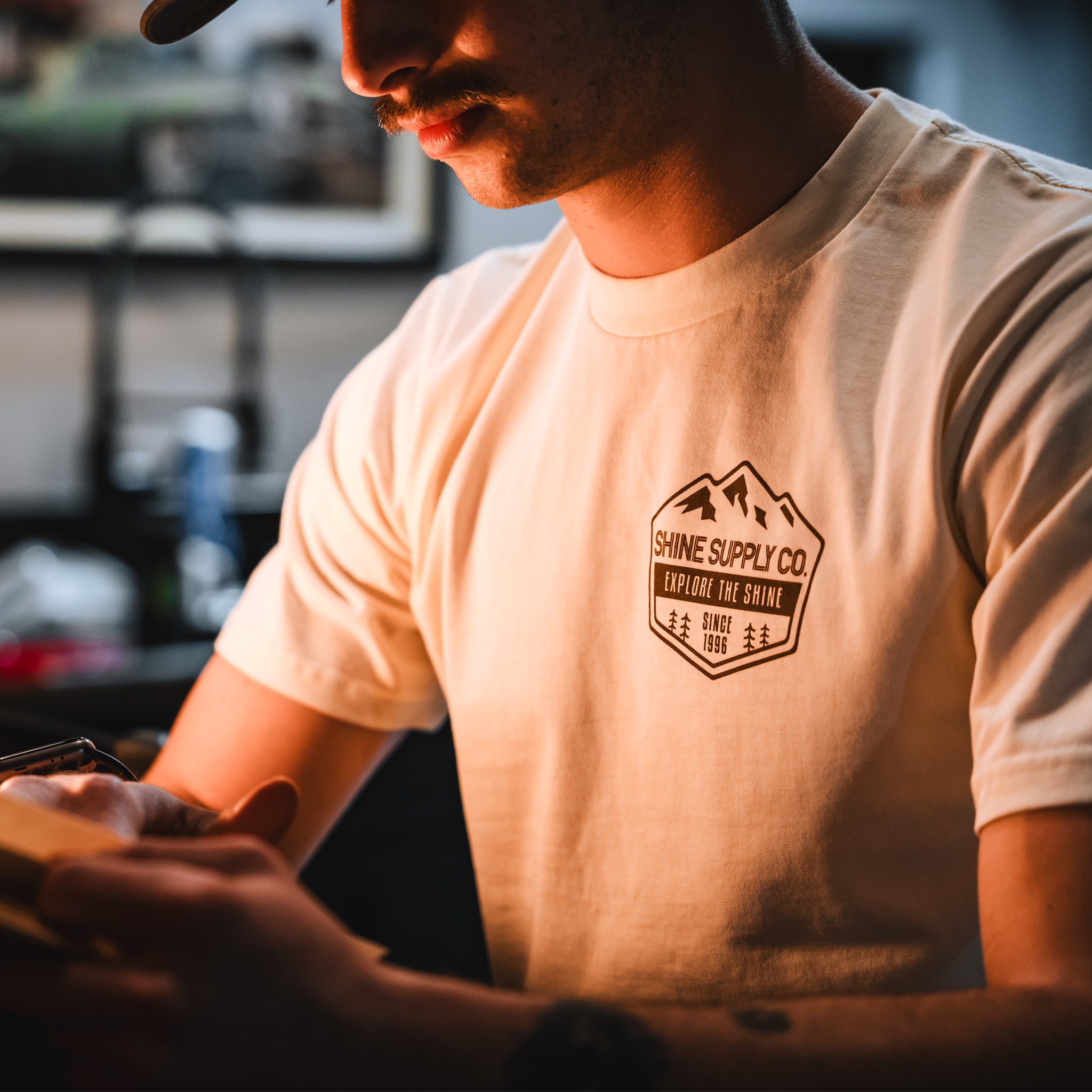 Man wearing ecru t-shirt with "Shine Supply Co." logo, engaging in a task.