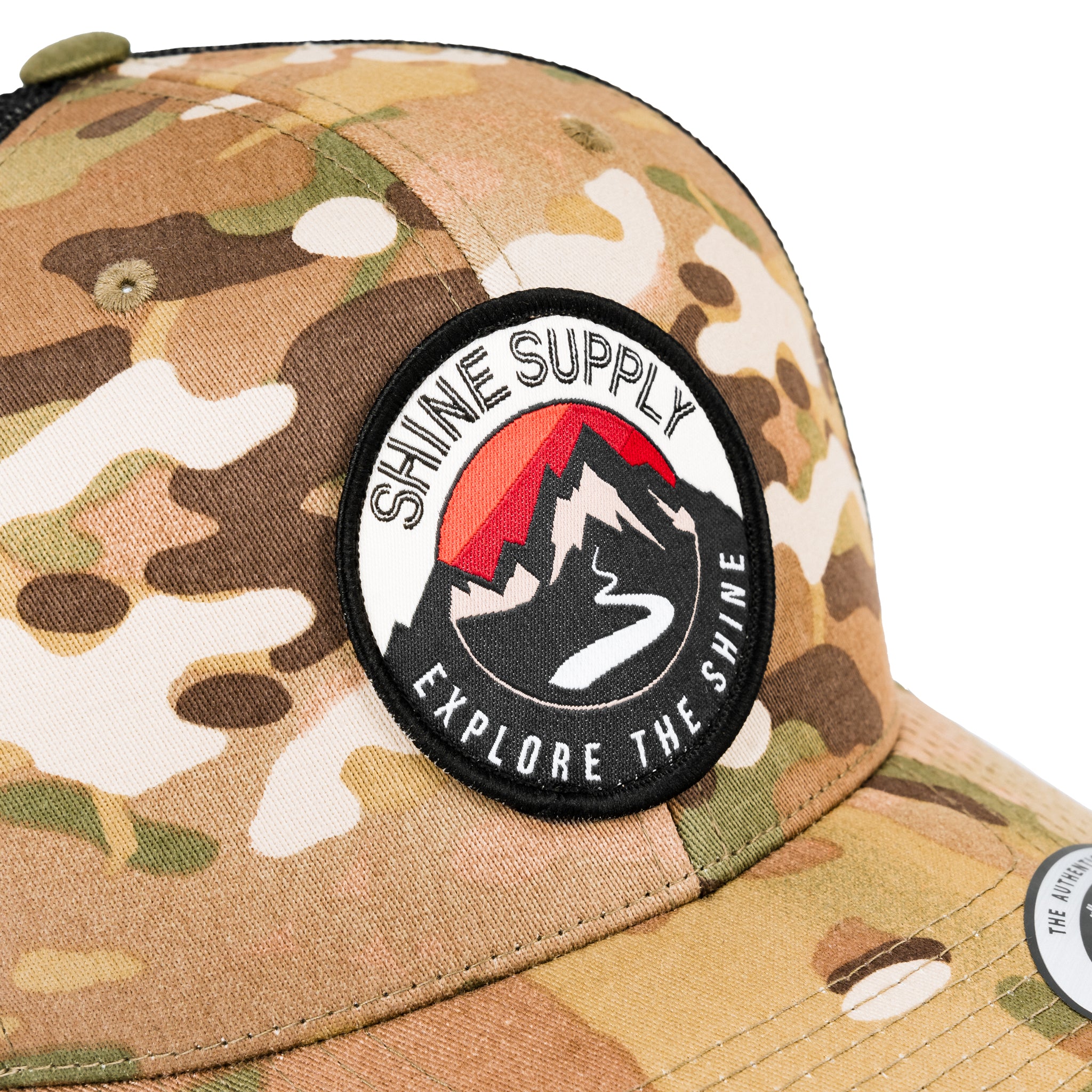 Close-up of a camo trucker snapback hat with a 'Shine Supply' logo patch featuring a mountain.