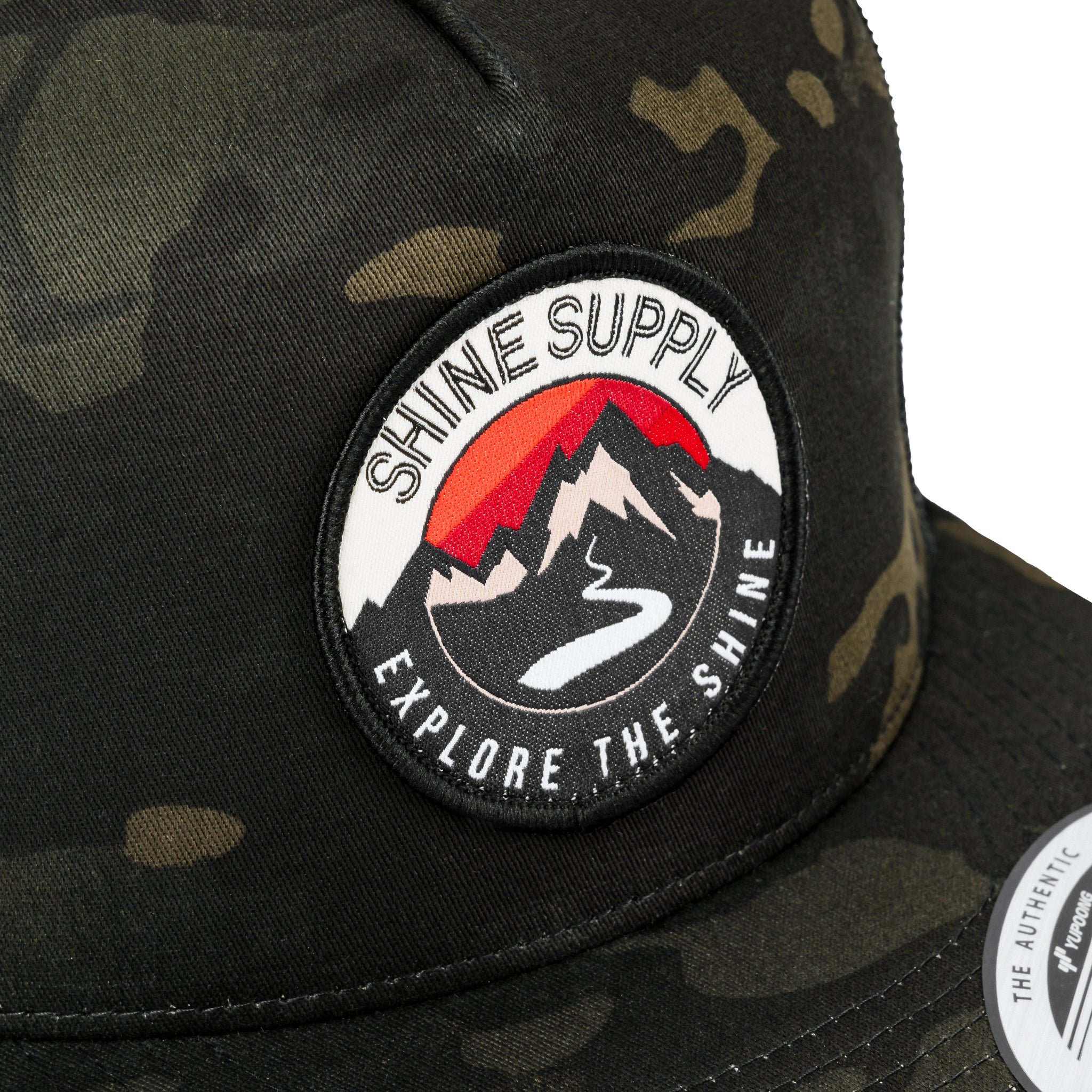 Close-up of a dark camo trucker snapback hat with a "Shine Supply Explore the Shine" logo patch