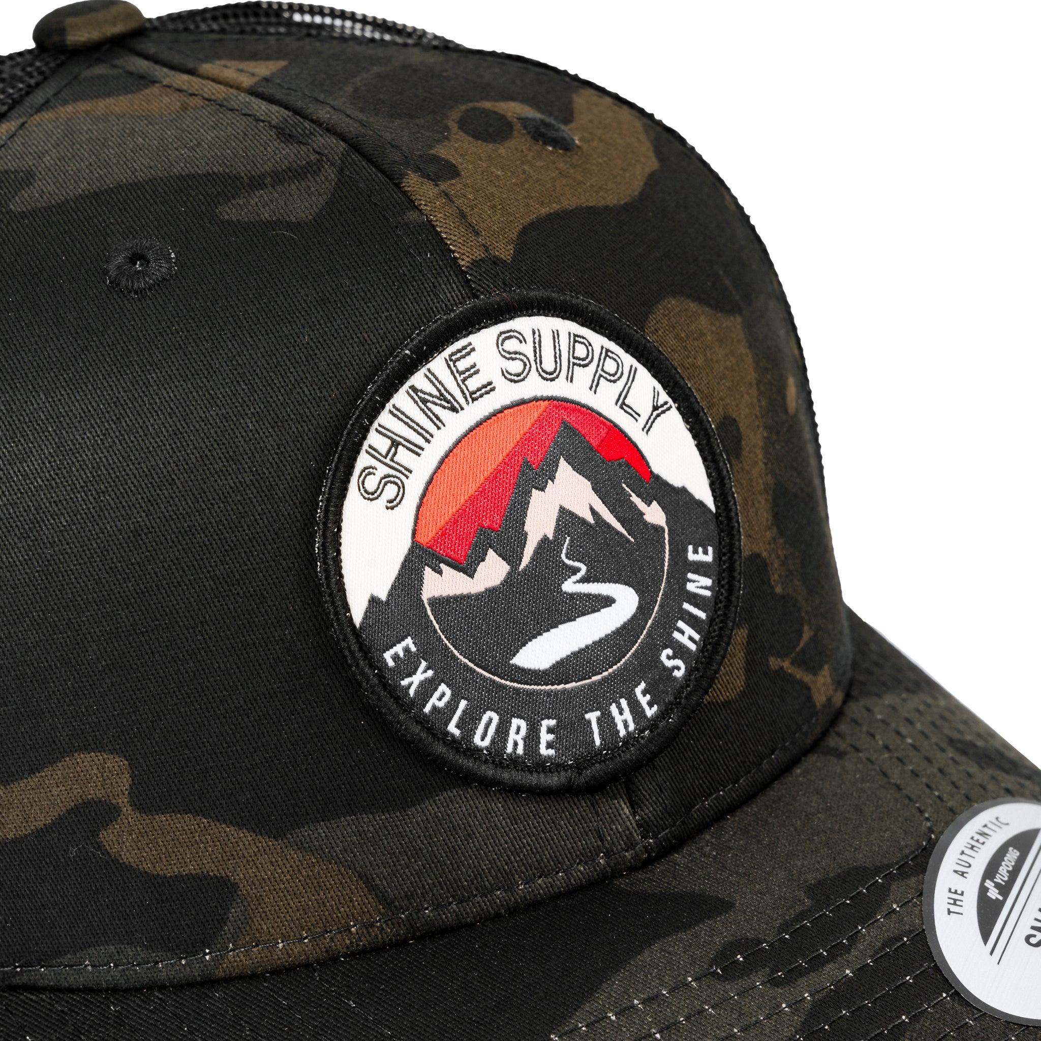 Close-up of a dark camo trucker snapback hat with a curved bill, featuring a "Shine Supply Explore The Shine" logo patch.