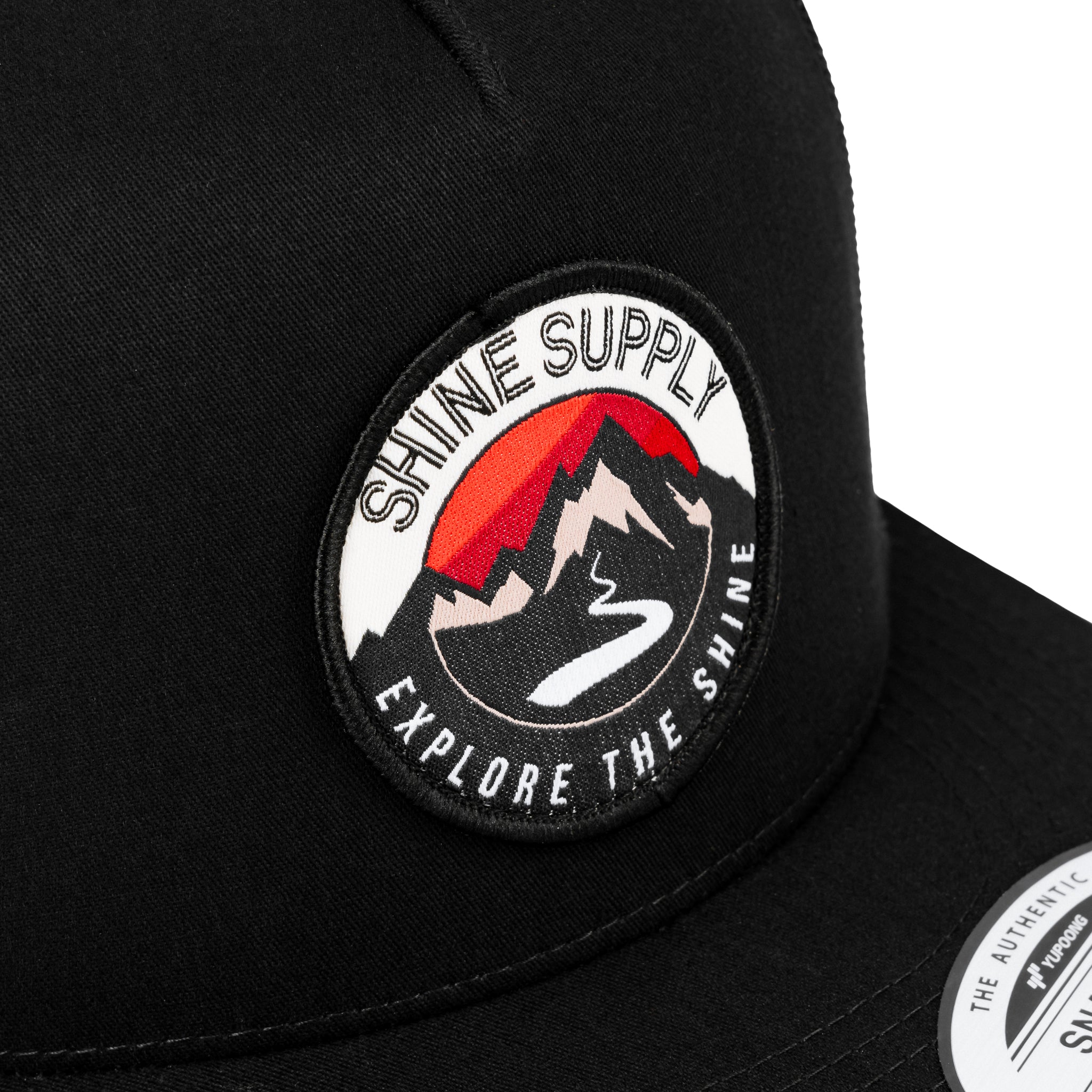 Close-up of a black trucker snapback with a "Shine Supply - Explore The Shine" logo patch.