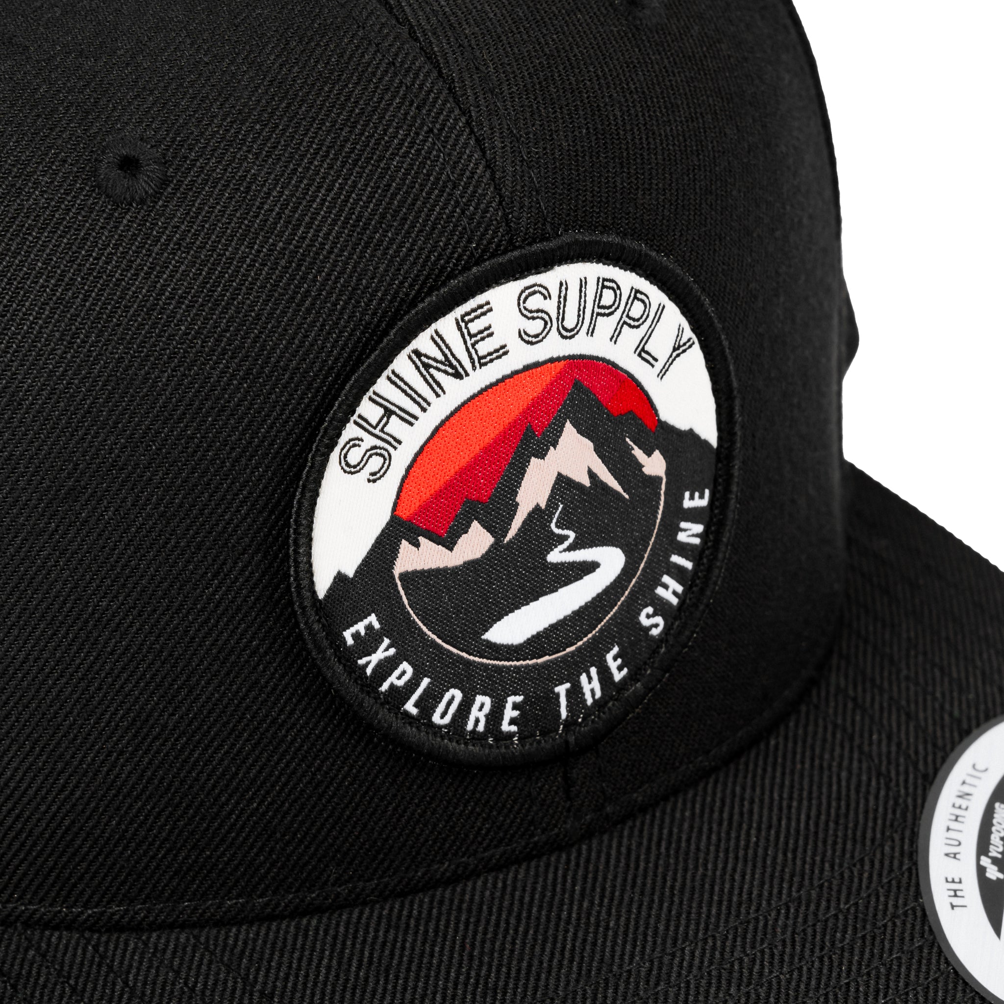 Close-up of a black snapback hat with "Shine Supply Explore the Shine" logo featuring a mountain design.