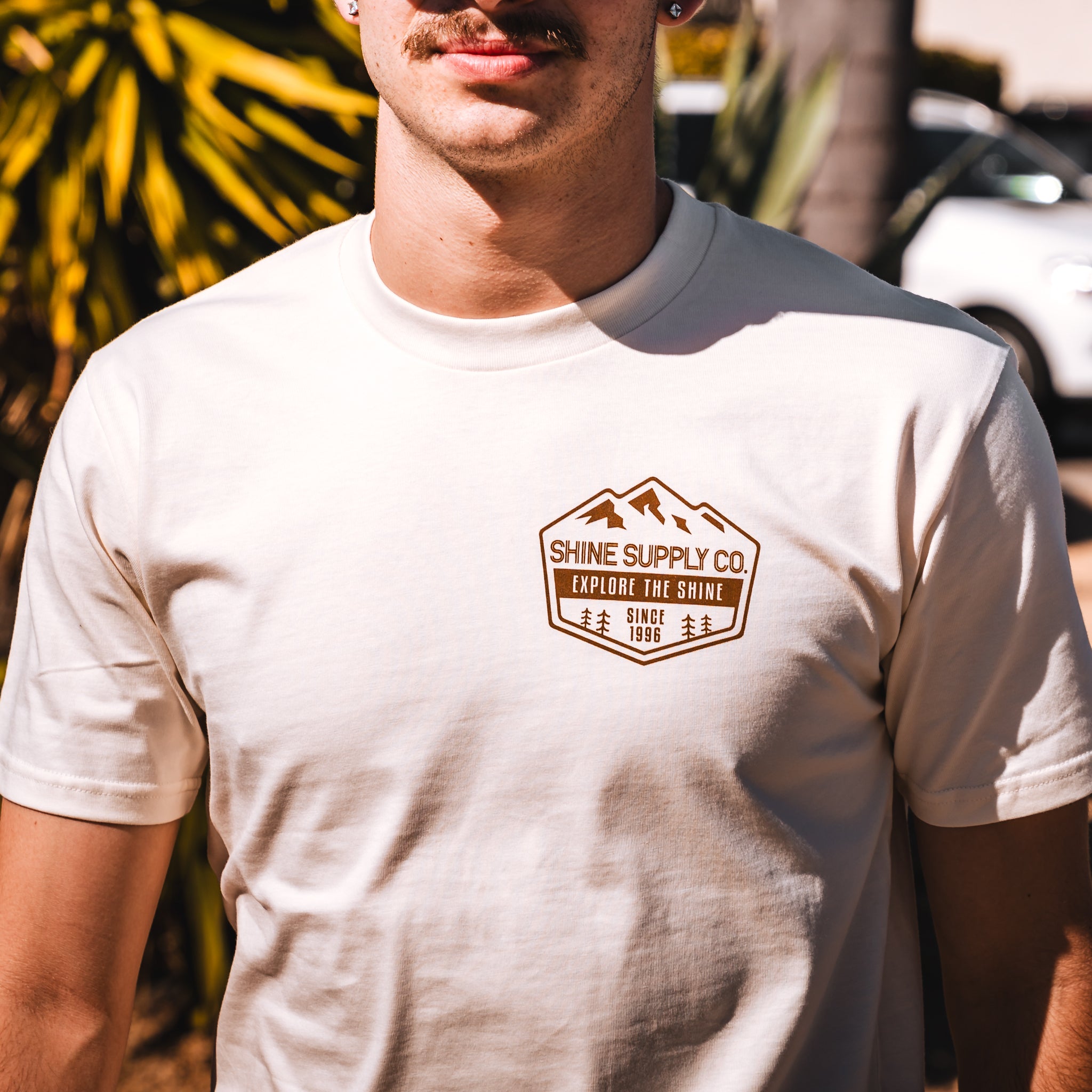 Man wearing ecru T-shirt with 'Shine Supply Co. Explore The Shine' logo in a sunny outdoor setting.