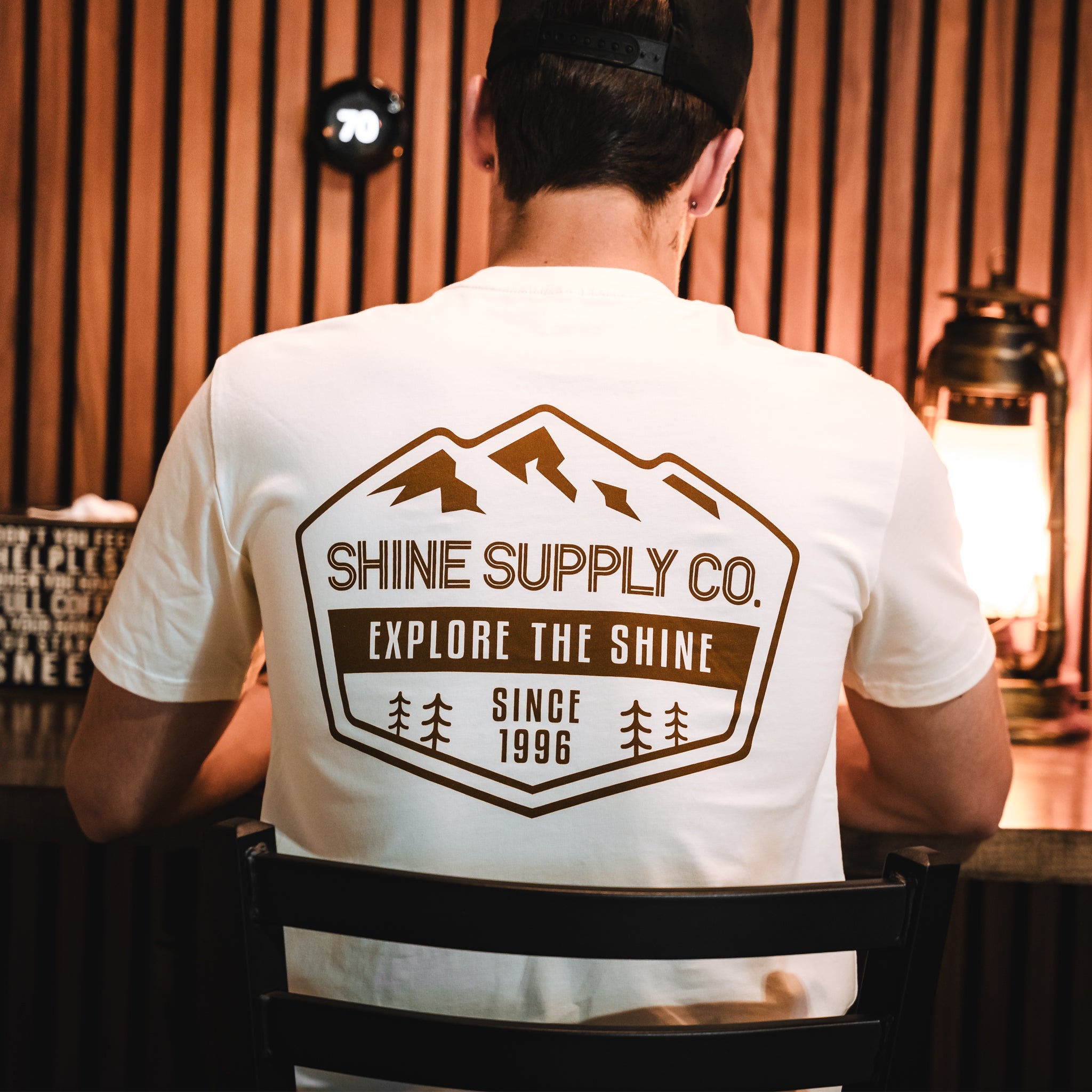 Man in ecru t-shirt with 'Shinie Supply Co. Explore The Shine Since 1996' logo, indoors.