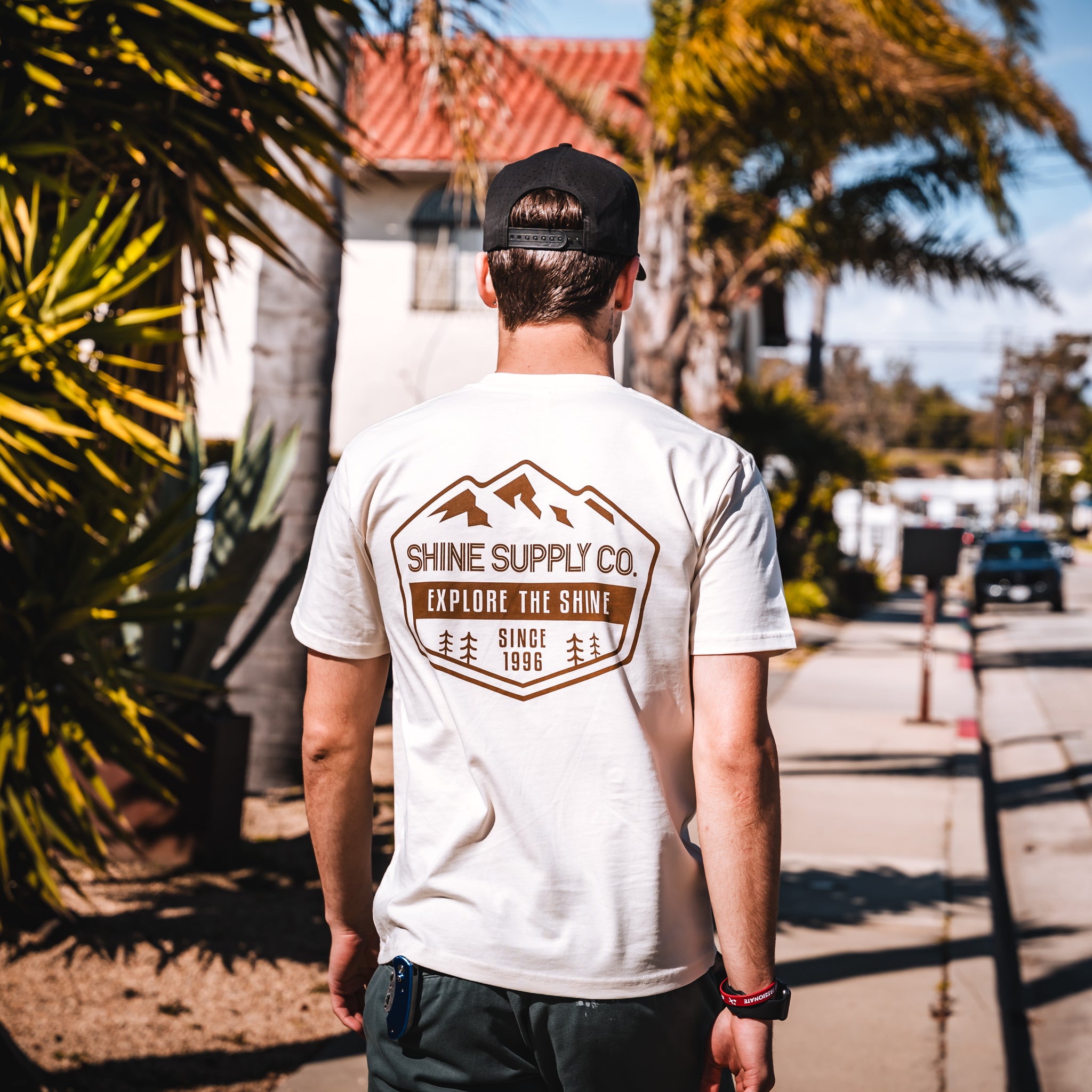 Man wearing an ecru t-shirt with 'Explore The Shine' graphic, walking on a sunny street.